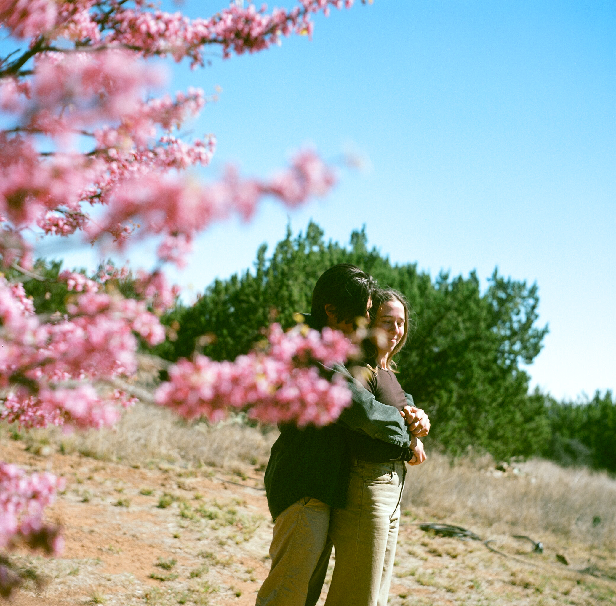 Henry embracing Katherine from behind with a cherry blossom in the foreground, taken in Fort Davis, West Texas, during their engagement session.”