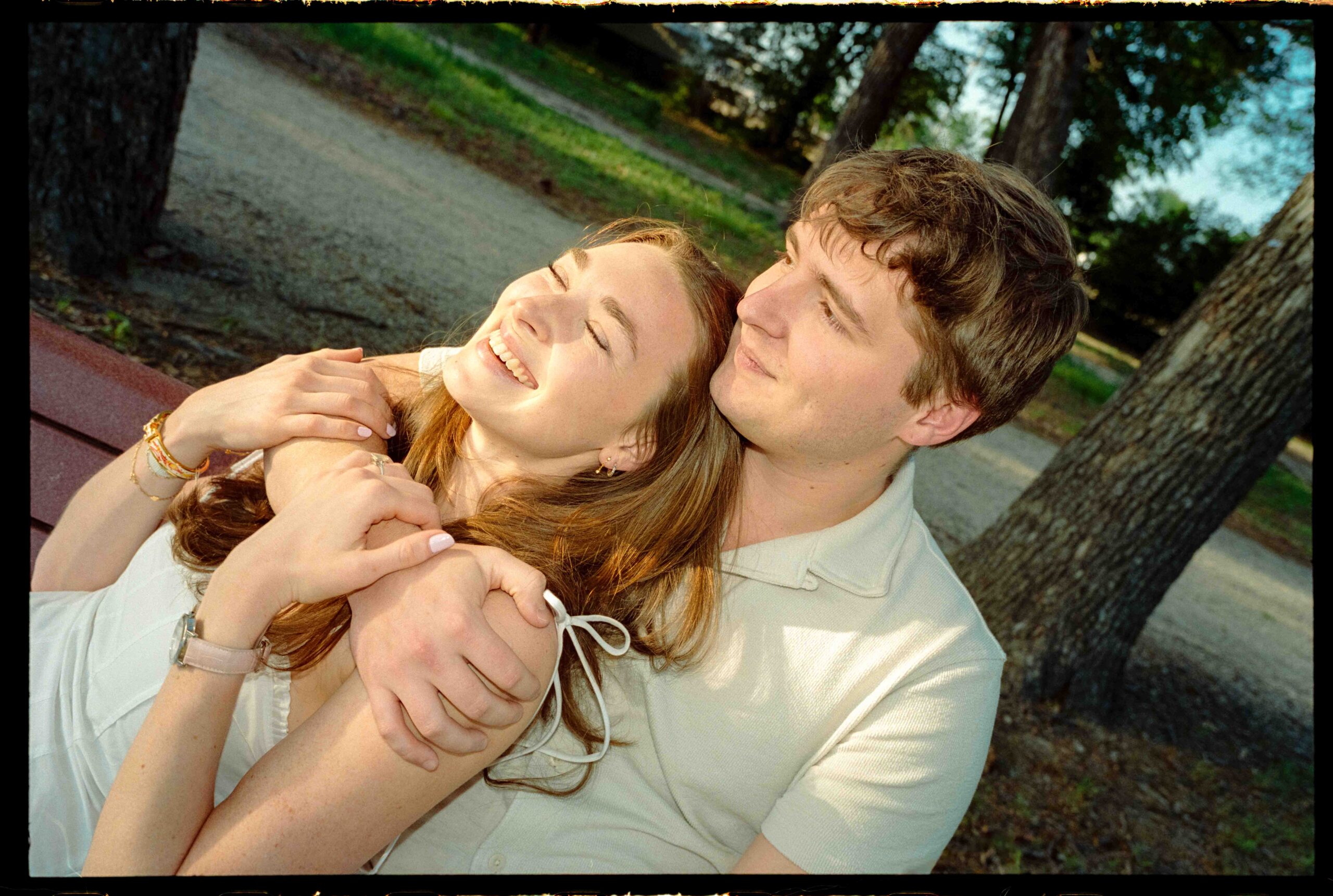 Claire and Gabe cuddling on a bench at the Trinity River during golden hour, smiling and holding each other with sunlight streaming through the trees.